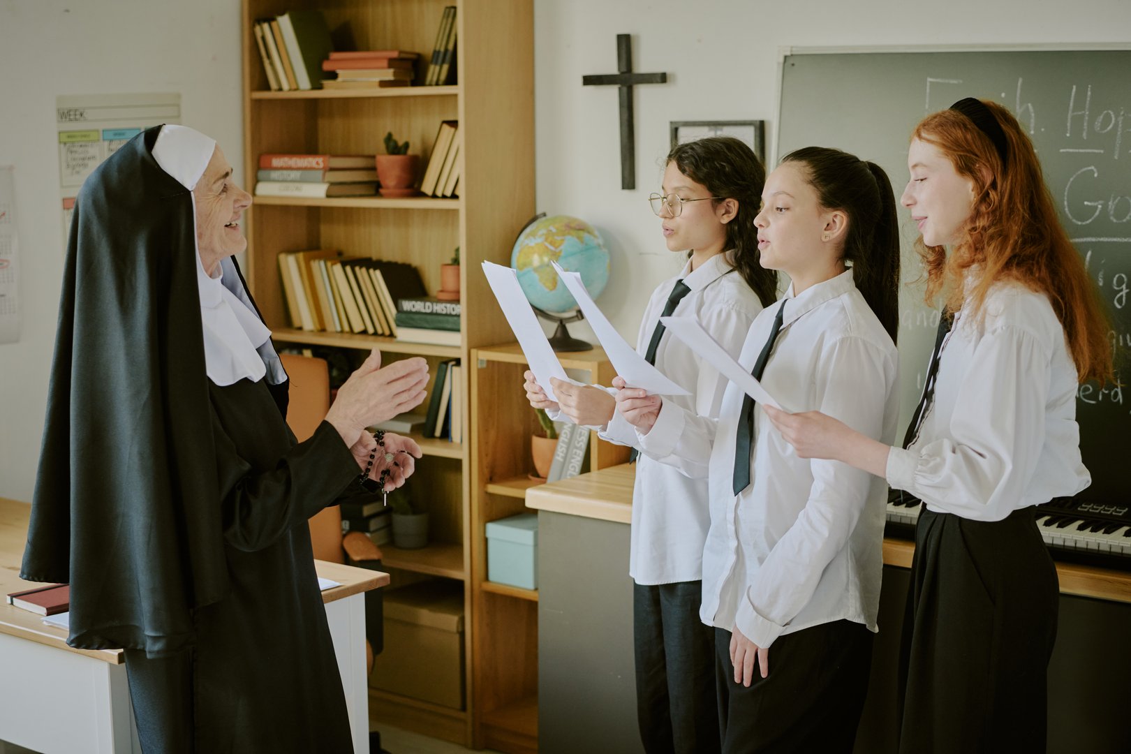Teacher wearing habit discussing notes with three students standing near classrooms blackboard and globe. Classroom contains bookshelves filled with books and other educational materials