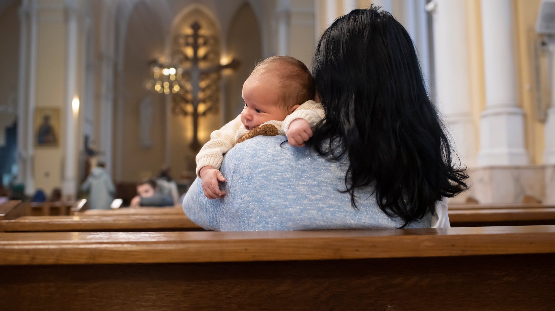 A woman holds a baby boy and sits on a bench in a Catholic church