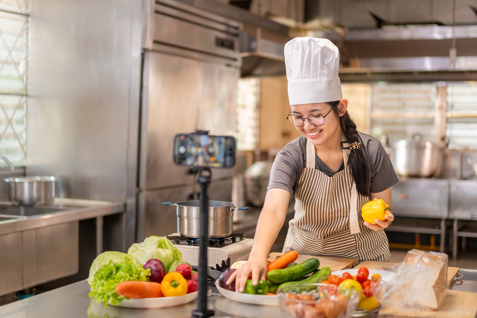 A smiling young adult Asian female chef records a cooking video on her phone in a professional kitchen, preparing fresh vegetables. Ideal for content creation, online tutorials, or culinary demonstrations.