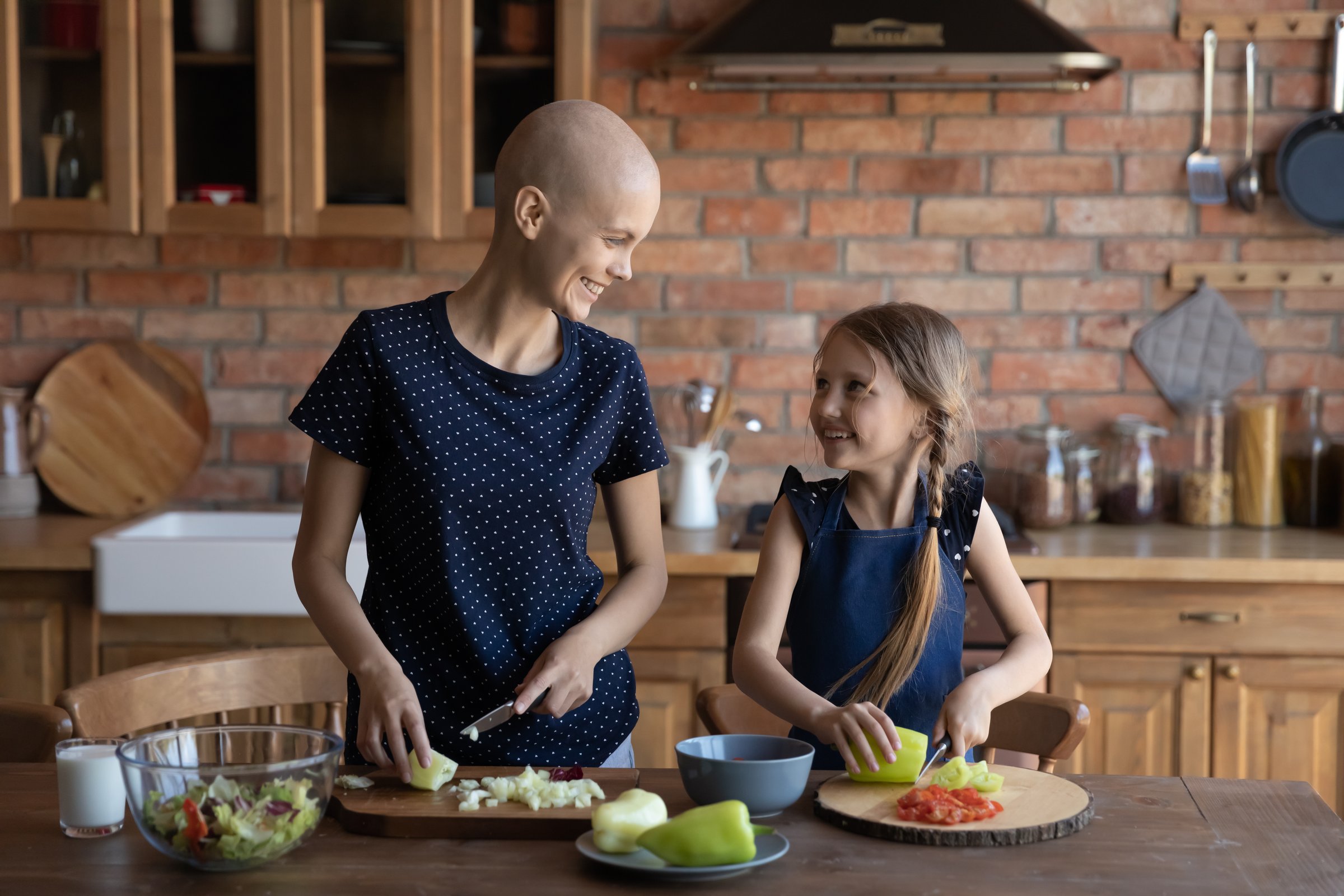 Happy sick hairless mother cancer patient and little daughter cooking together, chopping fresh vegetables for salad, standing at wooden kitchen table, unhealthy mum and adorable child girl having fun