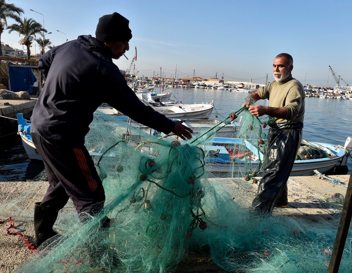 Two fishermen untangling nets by a dock with boats in the background on a clear day.