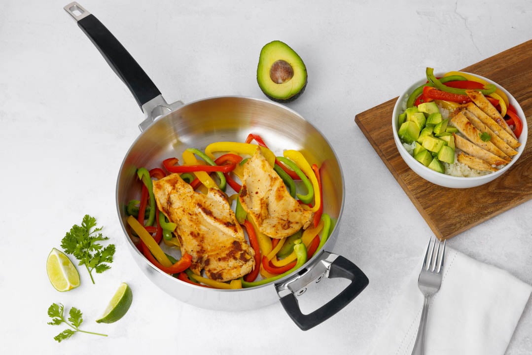 Cooked chicken breast with bell peppers in a pan, next to a bowl with chicken, avocado, and peppers, with lime and parsley.