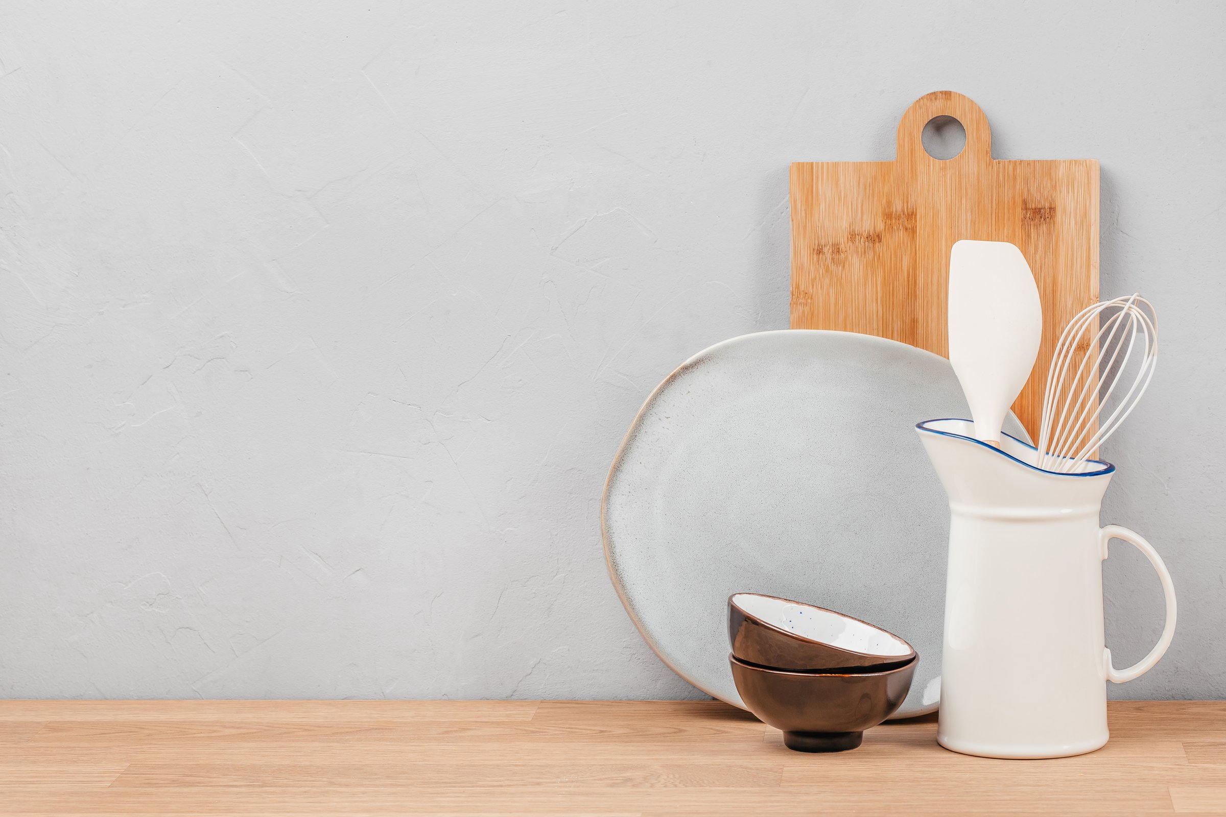 Ceramic kitchenware including plates, bowls, spatula, and whisk arranged with a wooden cutting board and pitcher on a light wooden surface against a textured wall background.