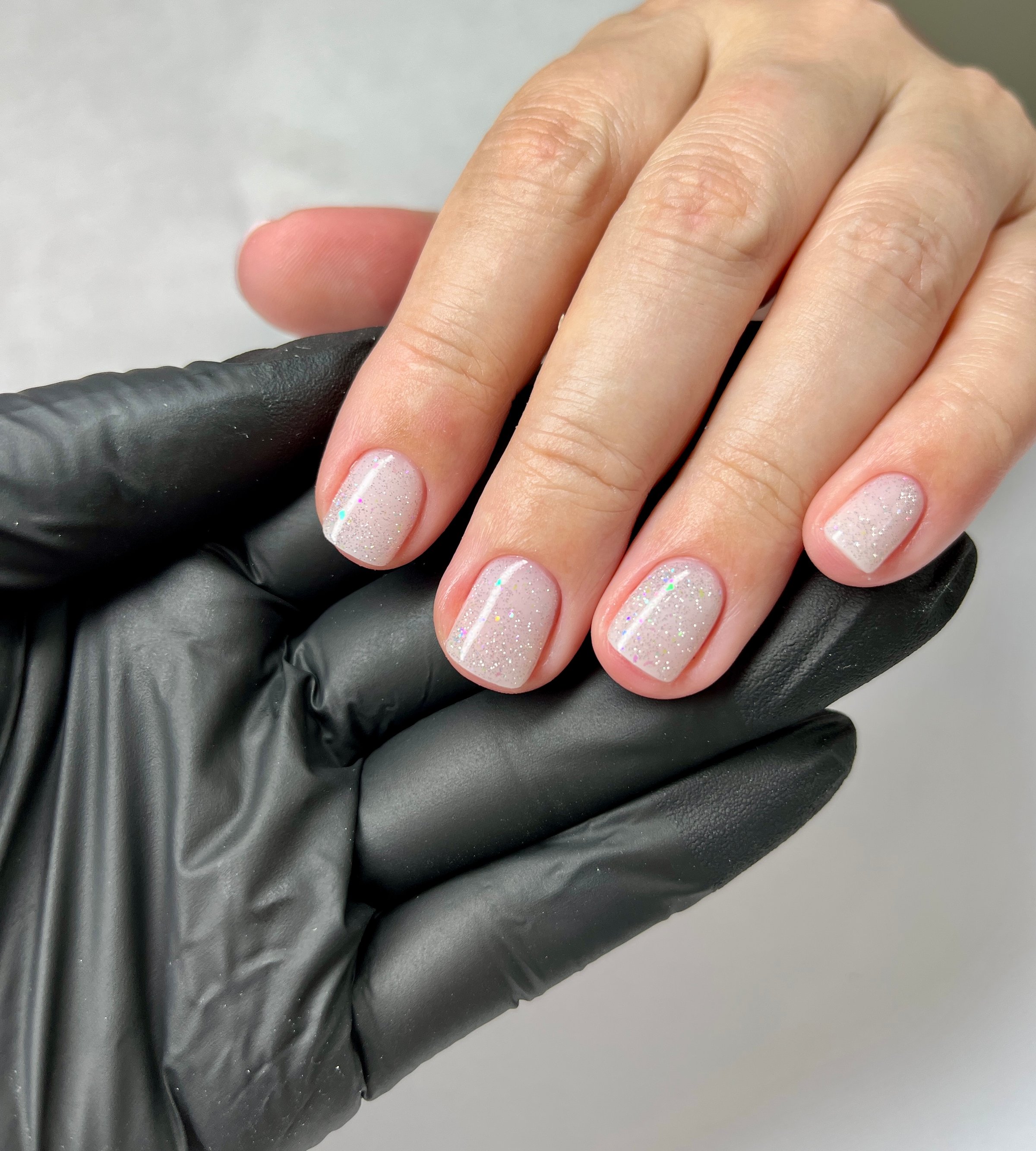 Closeup of a woman's hand with short glossy nude nails and glitter gel polish. Professional manicure in beauty salon. Hand in black glove holding client's fingers. Soft pink tone, minimalist and clean nail design. Perfect for advertising, cosmetology, hygiene, and spa care content.
