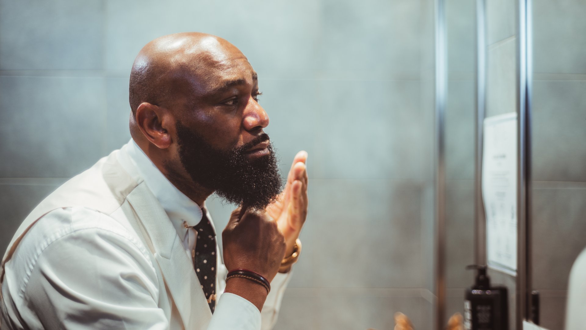 A profile view of a handsome bald black man grooming, brushing, and moisturizing the beard hair in front of the mirror in a bathroom of a luxury hotel; an African-American man taking care of his beard