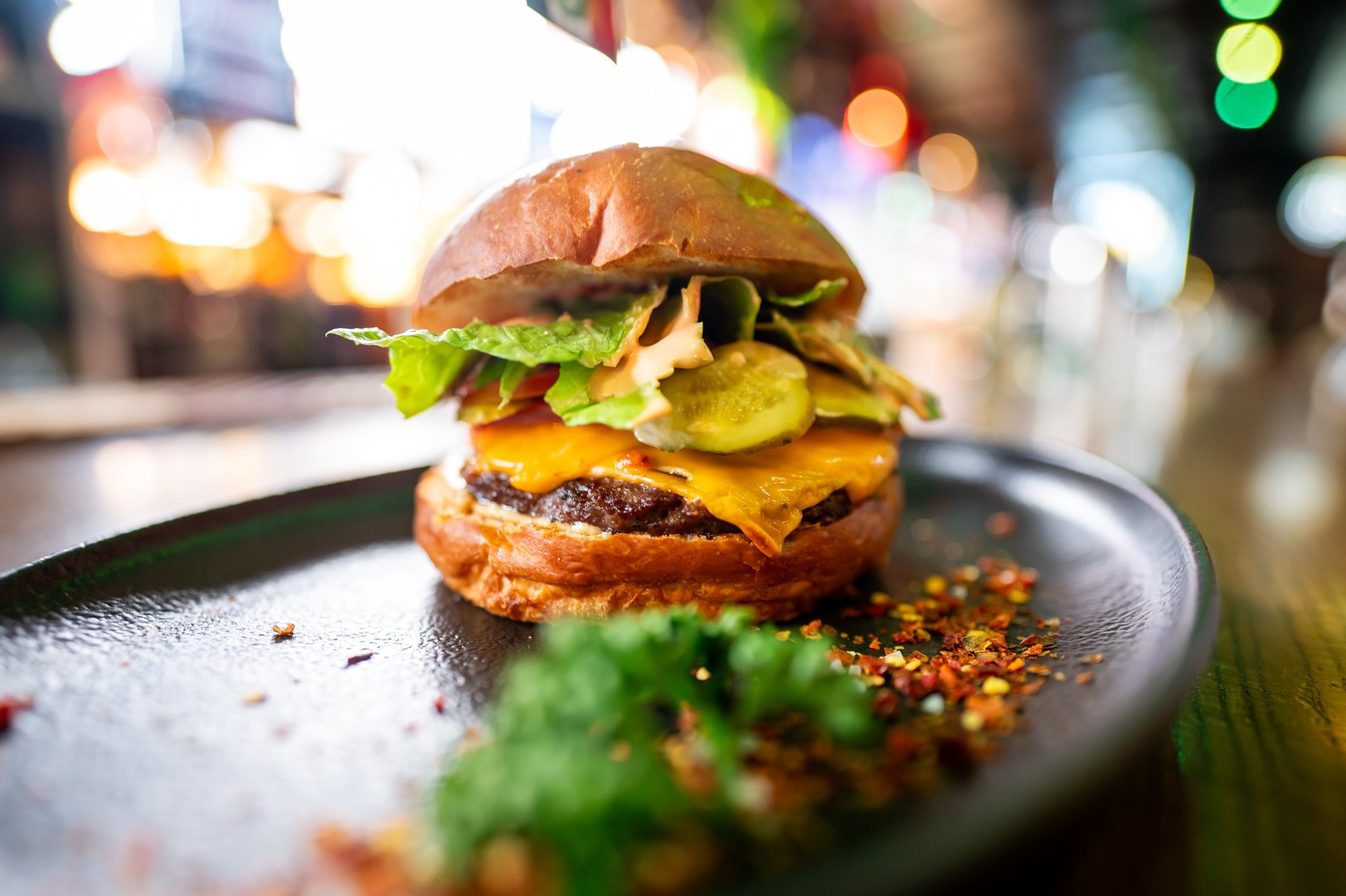A gourmet cheeseburger with a juicy beef patty, melted cheddar, lettuce, pickles, and sauce on a black plate. The background features colorful bokeh lights, creating a appetizing food presentation