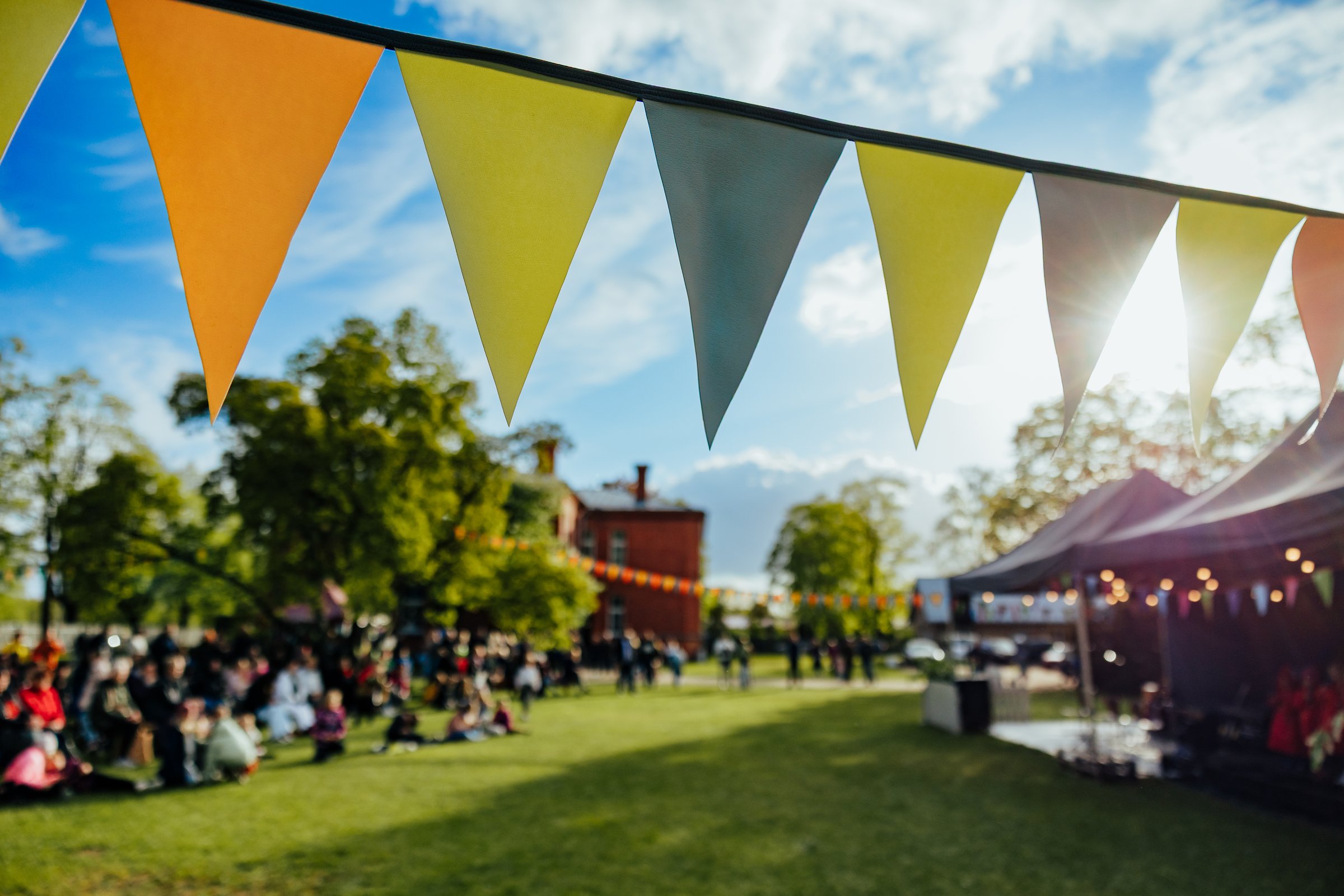 A lively outdoor festival scene with colorful bunting flags strung across a sunlit park, where people gather and enjoy a communal celebration under clear skies