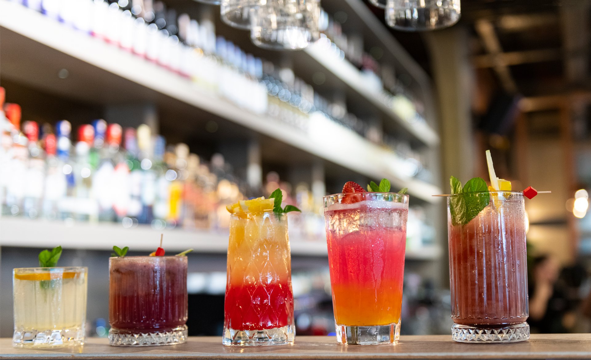 Five vibrant, colorful cocktails lined up on a sleek bar counter