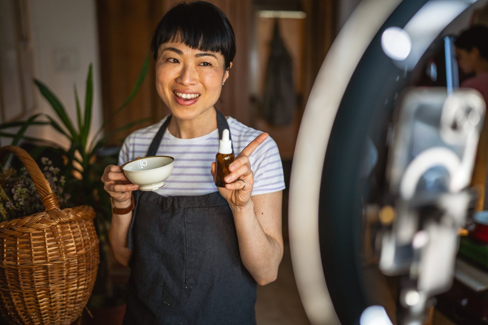 Mature japanese woman use mobile phone and ring light setting present dropper bottle nature serum at herbal workshop