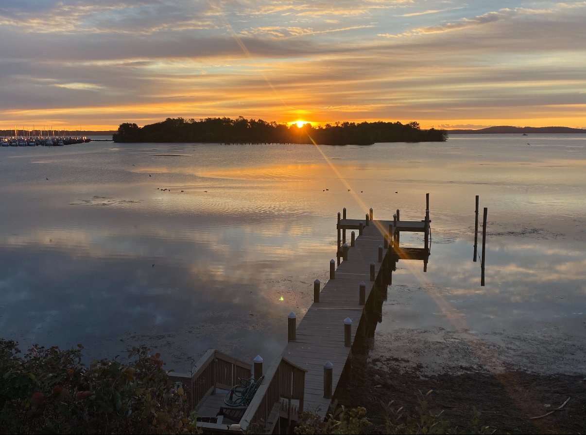 Sunset over a calm lake with a wooden pier, small island, and scattered clouds reflecting in the water.