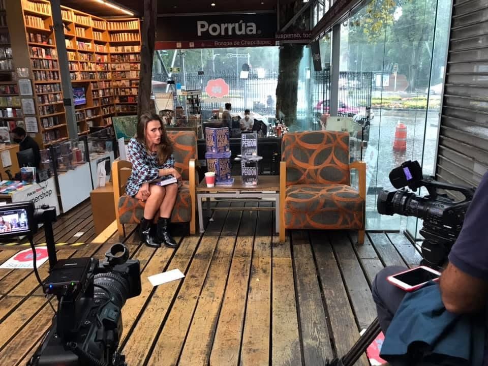 A woman sits next to a table with books in a bookstore setup, with cameras pointing towards her, suggesting an interview setting.