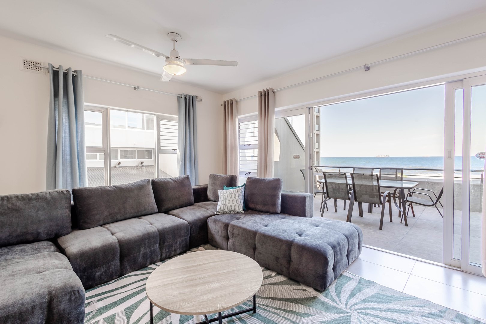 Modern living room with gray sectional sofa, round coffee table, and ocean view from a balcony with patio furniture.