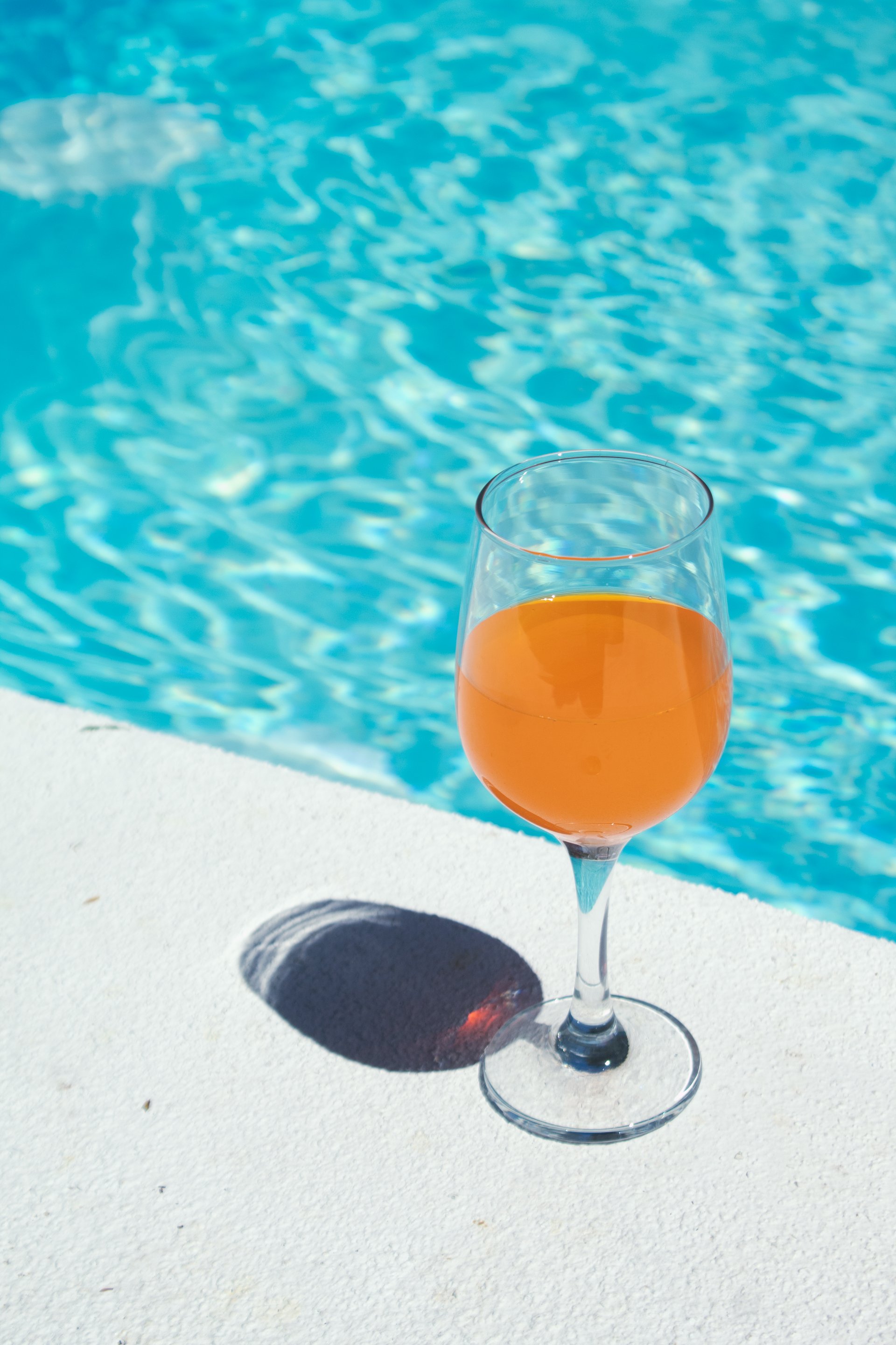 A wine glass filled with orange drink placed on the edge of a pool, casting a shadow on the white surface.