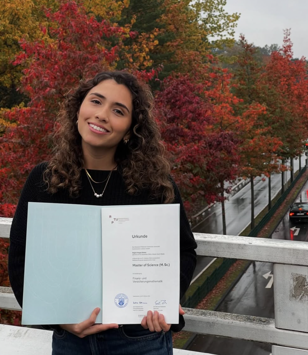 Person holding a master's degree certificate, standing on a bridge with autumn trees in the background.