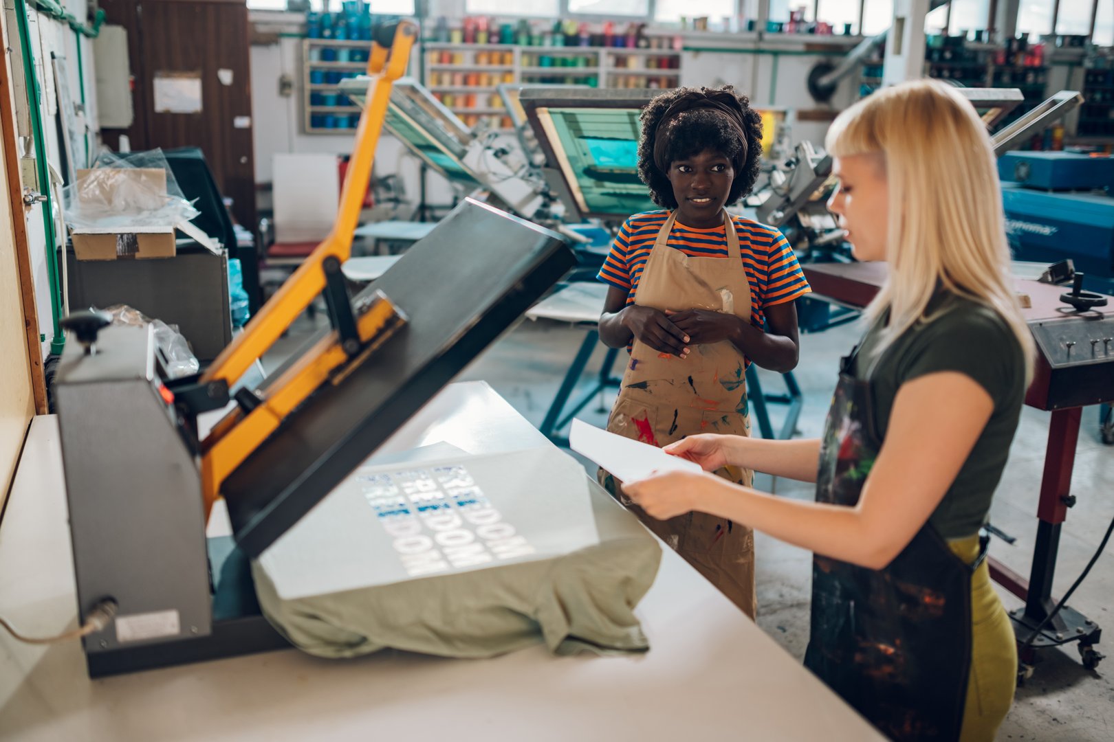 Portrait of a two multicultural female printing industry experts standing near heating press and working with silk screen printed t-shirt. Blond worker explaining process to her interracial colleague.