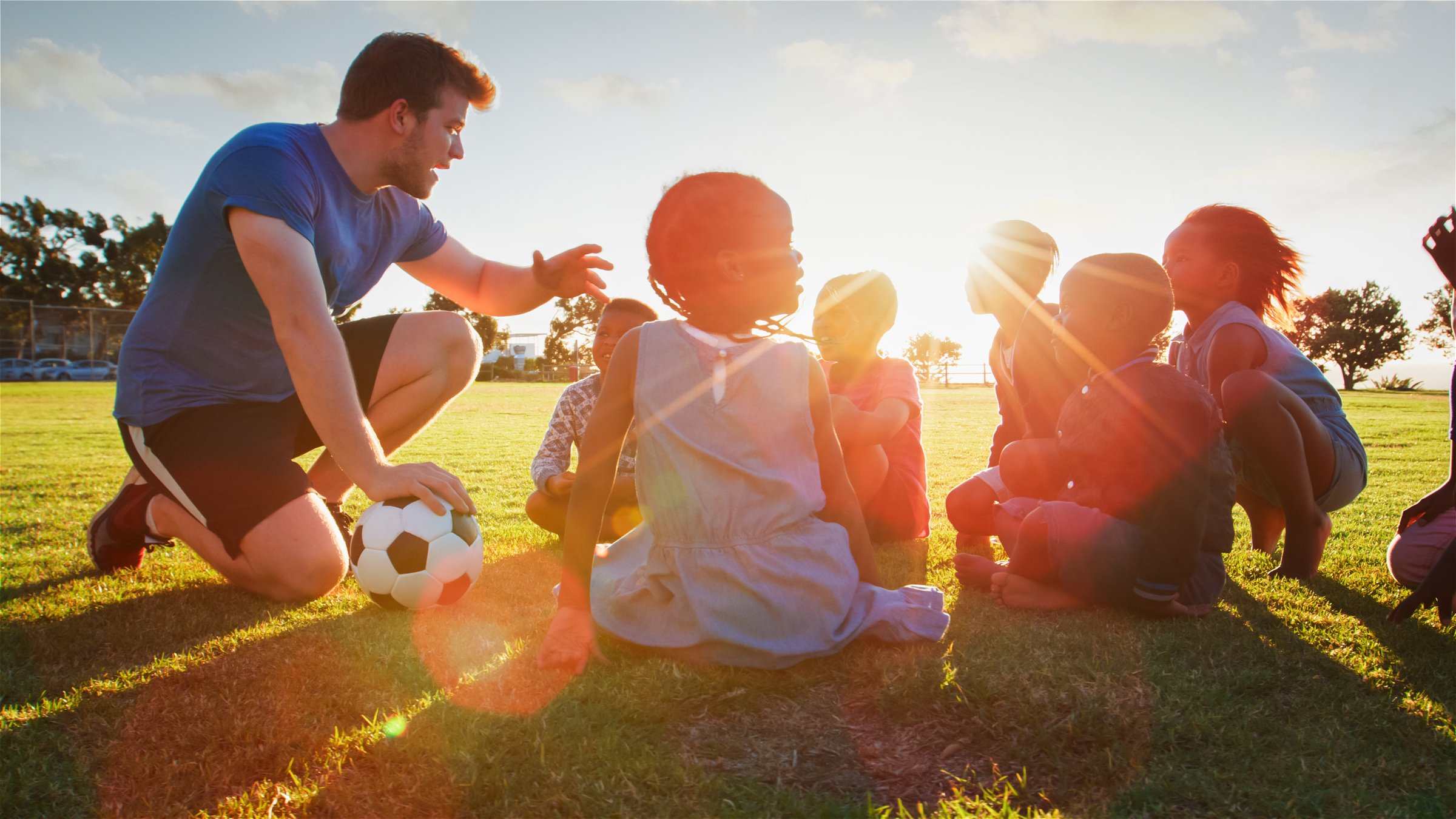 Coach teaches young athletes on a grassy field as the sun sets.
