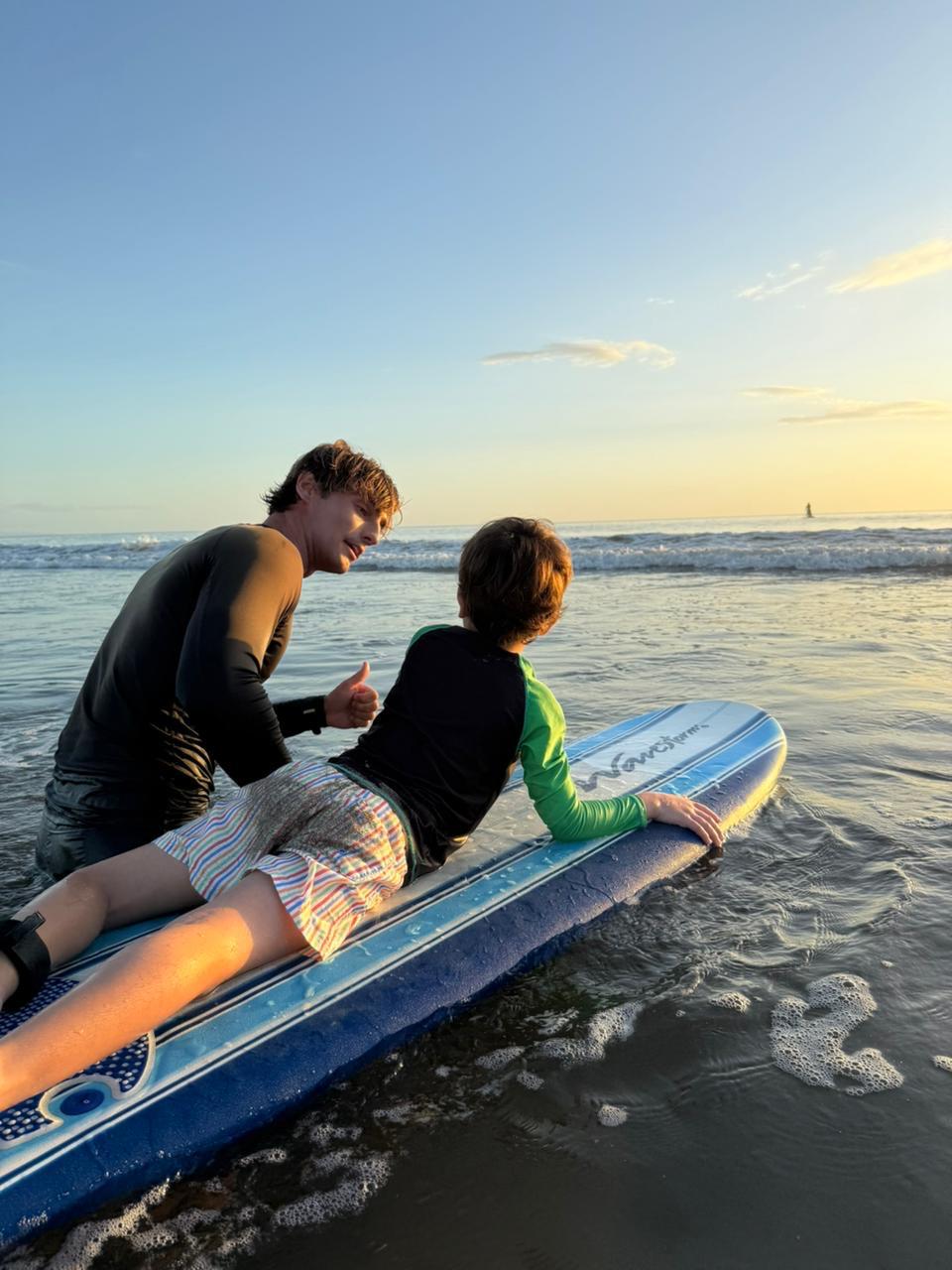 An adult teaches a child to surf at the beach during sunset. They are both on a surfboard facing the sea.
