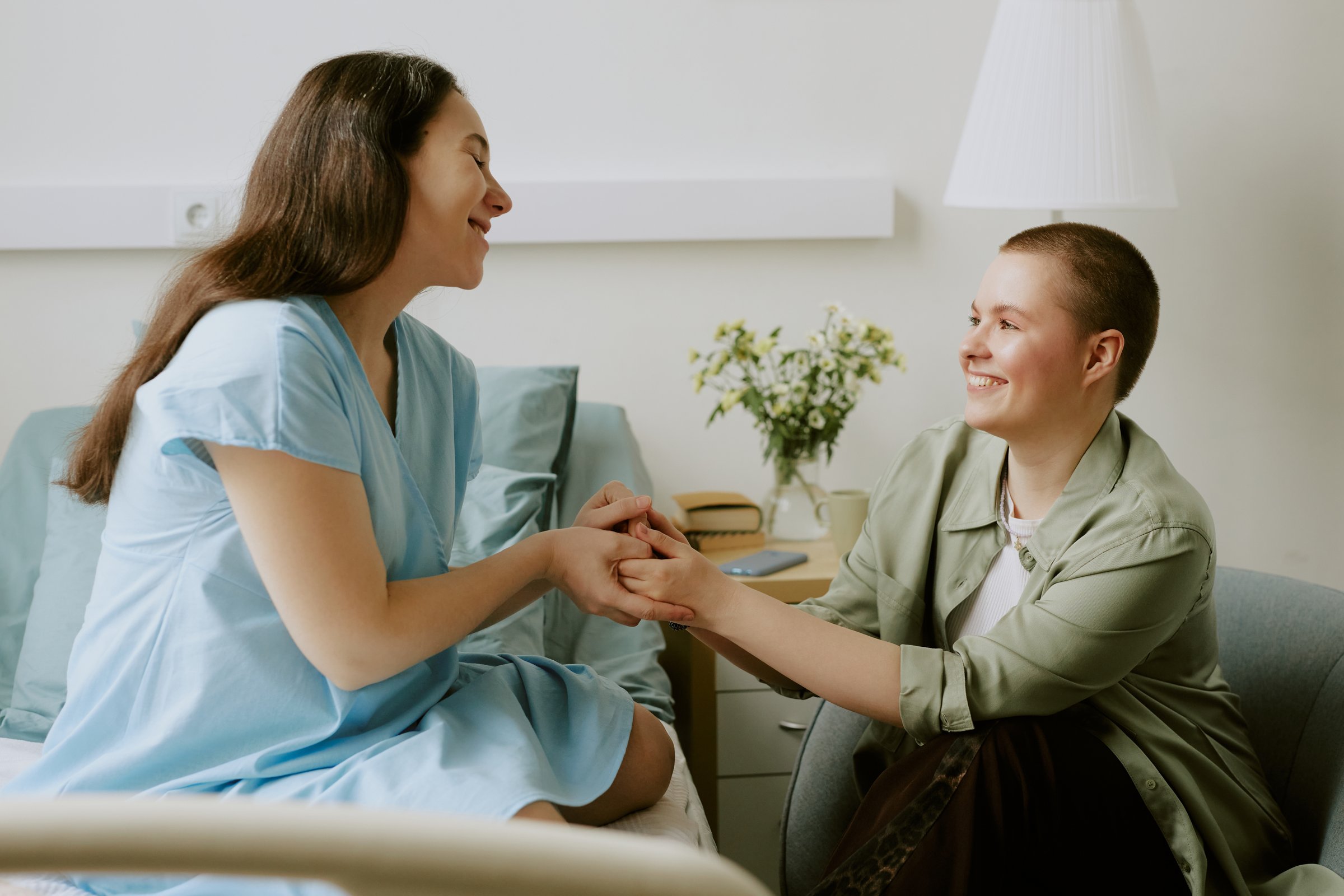 Pregnant patient sitting on bed and holding lovely her friends hands while they having lively talk