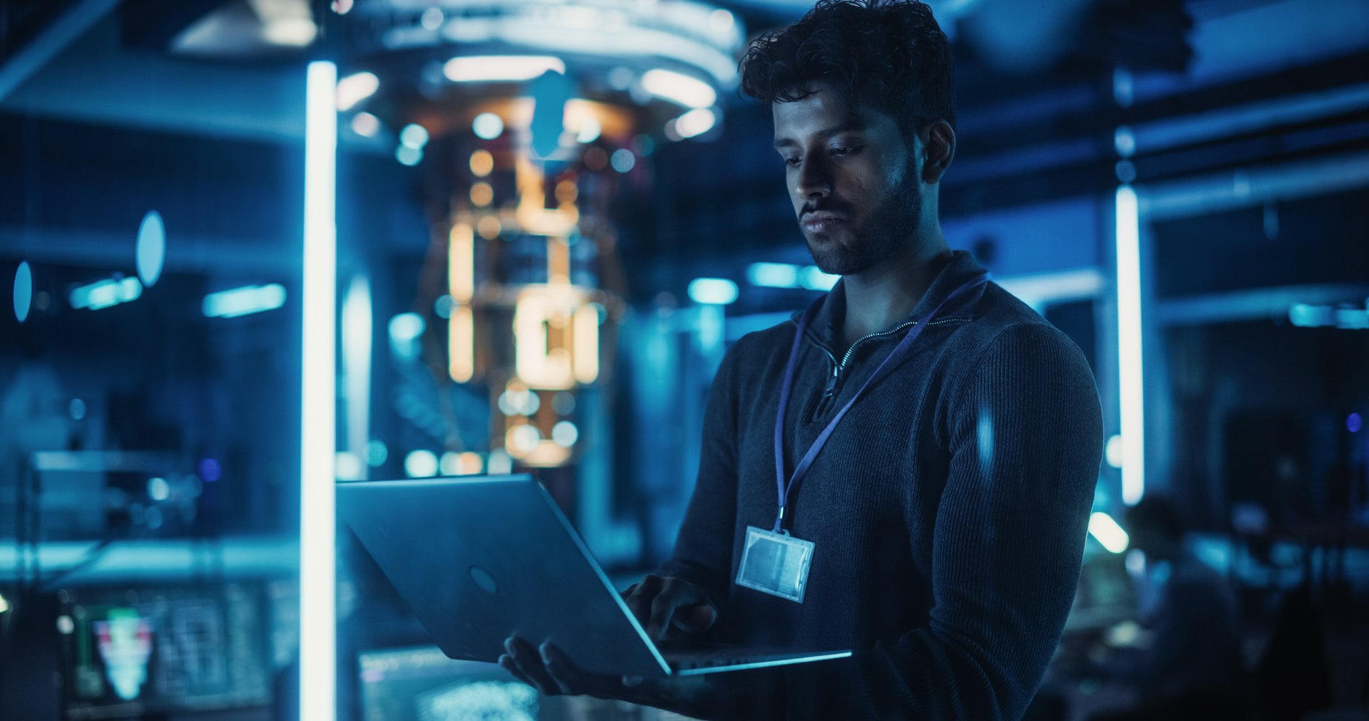 Indian Software Developer Standing in a Dark Modern Office, Working on a Laptop Computer. Young Diverse Specialist in Artificial Intelligence is Developing a Quantum Machine Learning Algorithm
