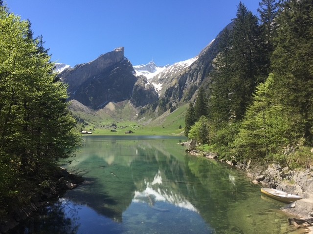 Alpine lake with clear water reflecting snowy mountains, surrounded by trees and a small boat on the right side.