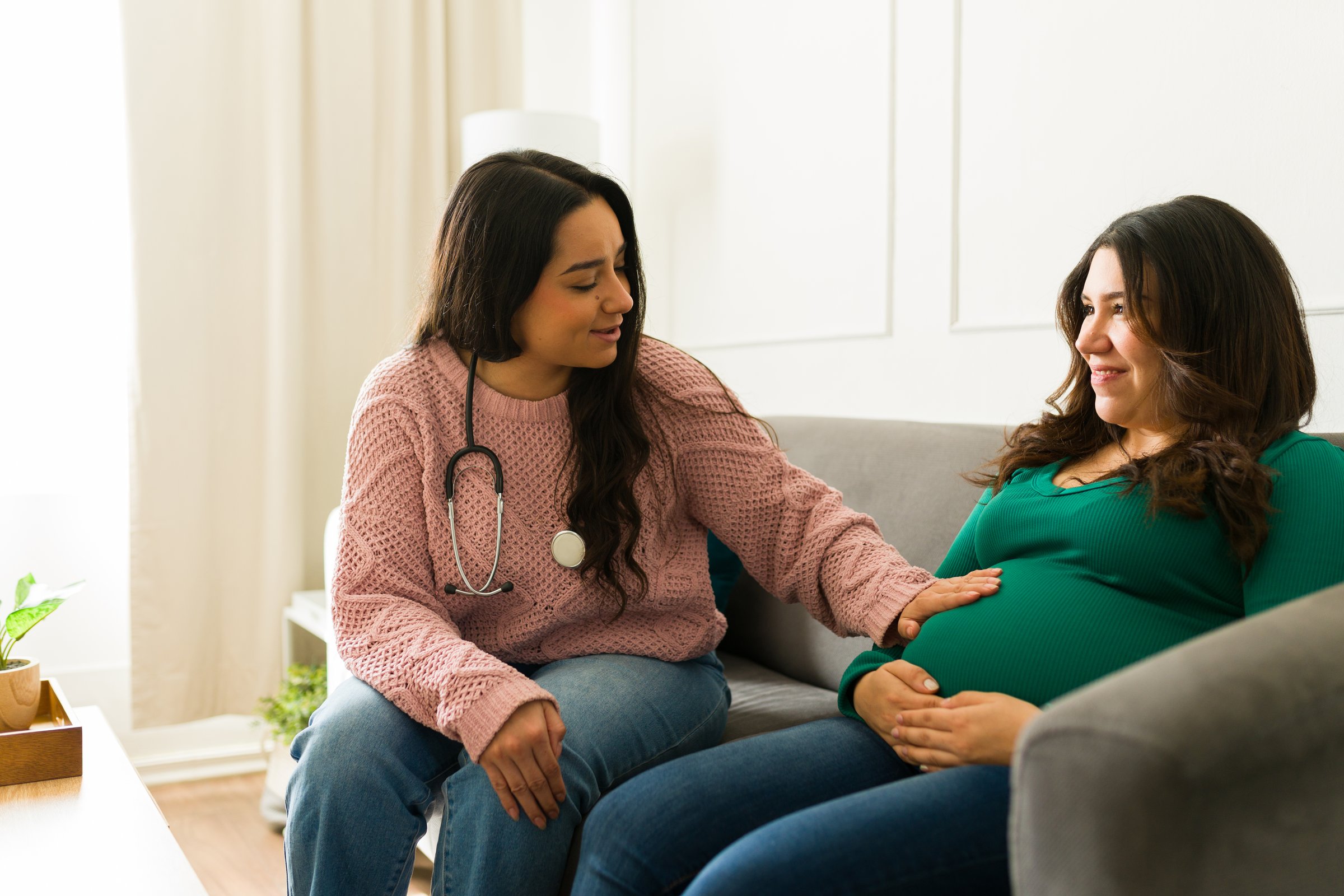 Female medical professional palpating expectant mother's abdomen during home consultation, offering specialized prenatal care and individualized support
