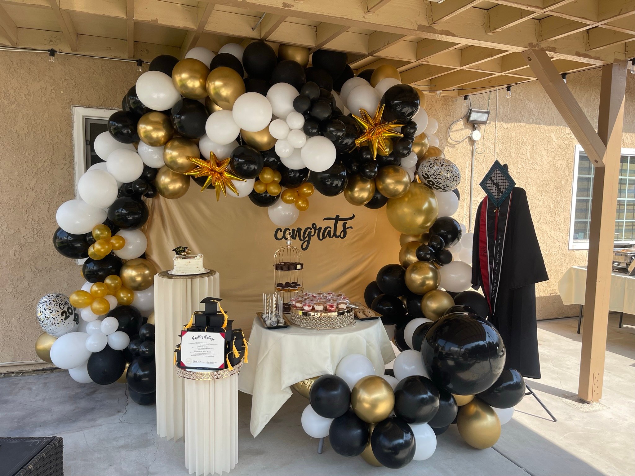 Graduation party setup with black, gold, and white balloons, a "congrats" sign, cake, cupcakes, and a graduation gown on display.