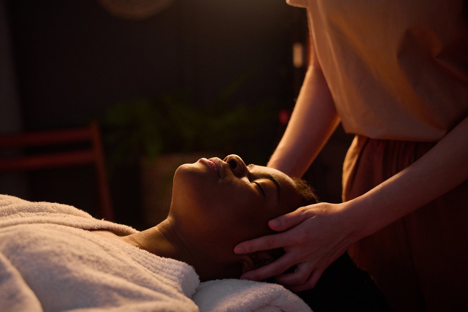 Medium close up of African American woman in bathrobe lying with closed eyes enjoying head massage by professional masseuse