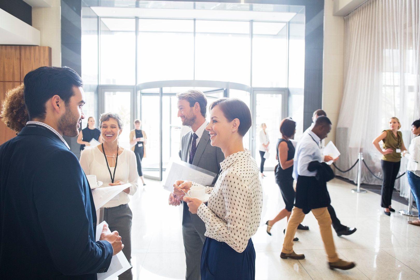 Professionals networking in a bright, modern lobby. People are conversing, holding papers, and walking about.