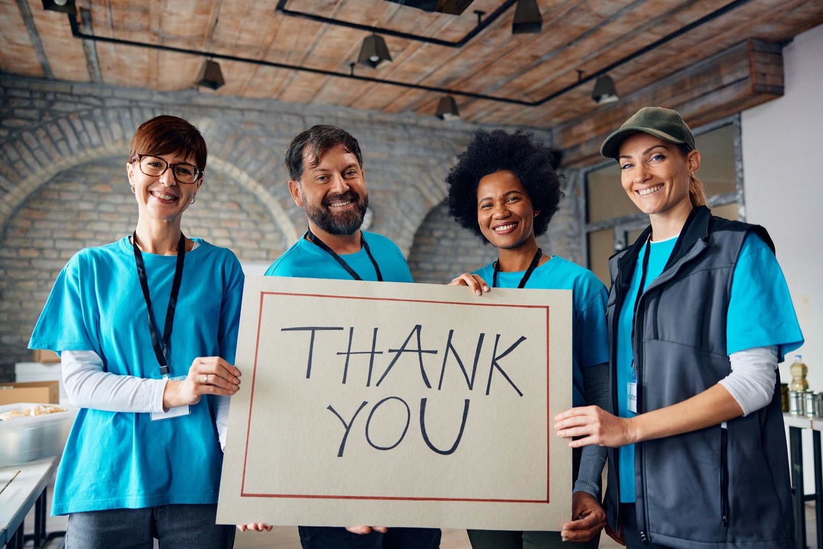 Happy team of volunteers with 'Thank you' placard and donation center looking at camera.