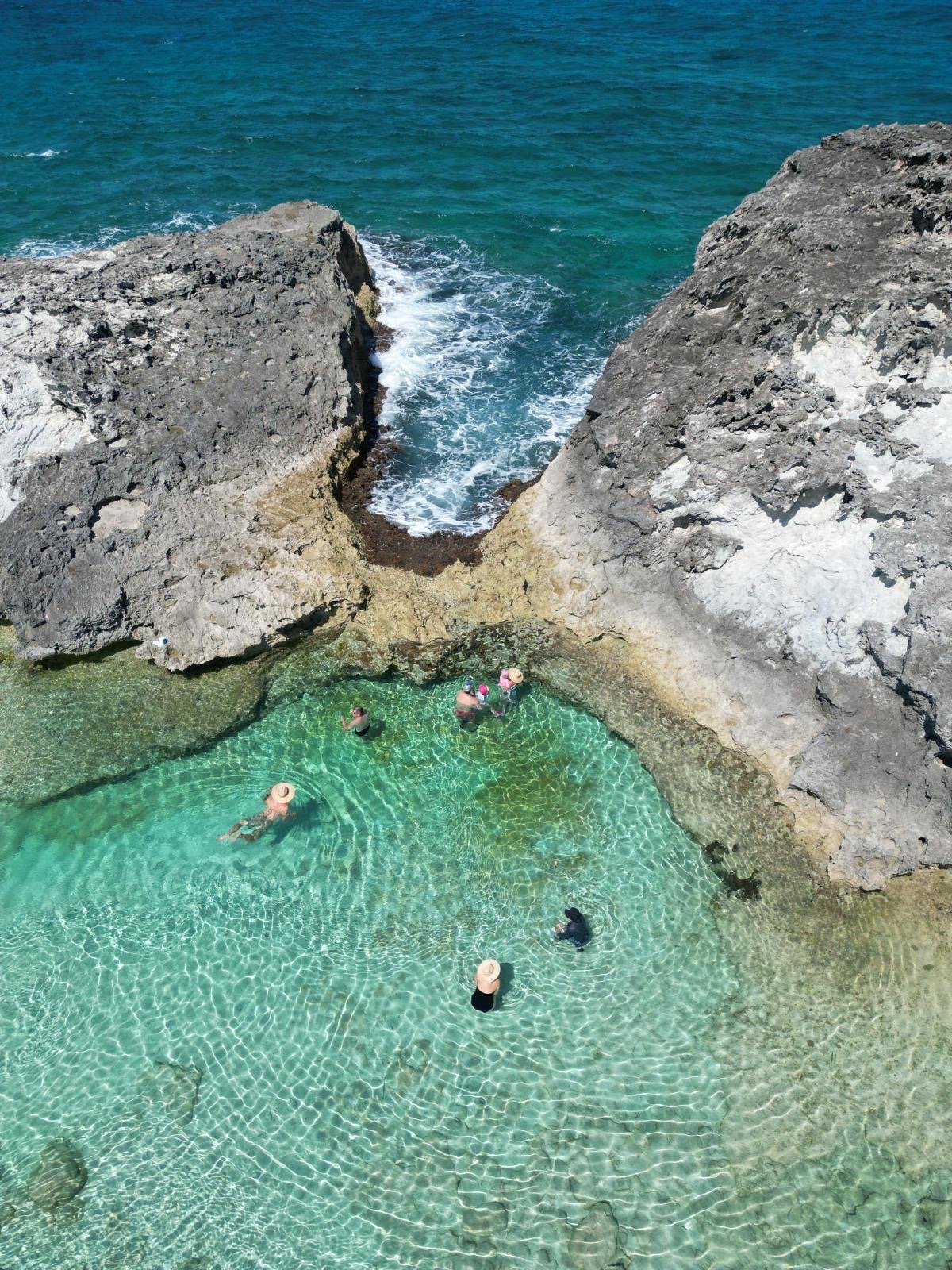 People swimming in a rocky coastal tidal pool surrounded by clear turquoise water with waves crashing nearby.