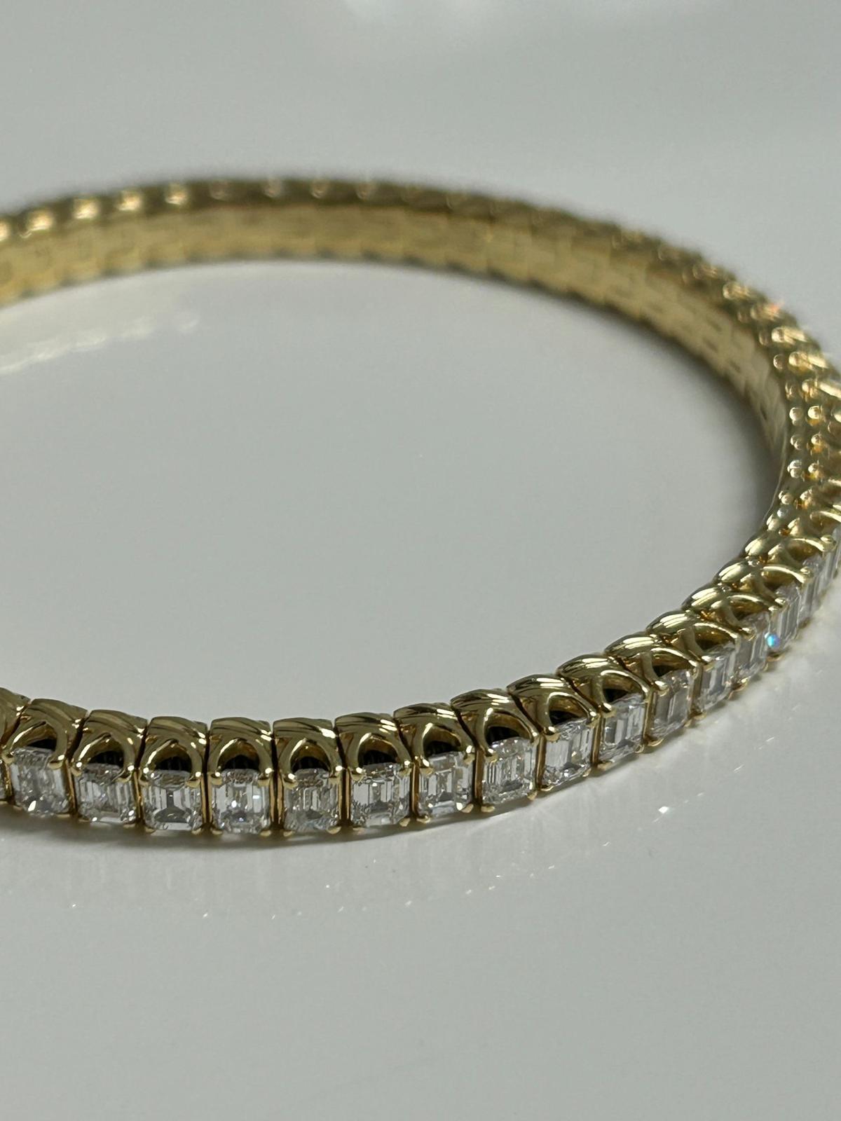 A gold bracelet with a row of rectangular clear gemstones set in individual settings, displayed on a reflective surface.