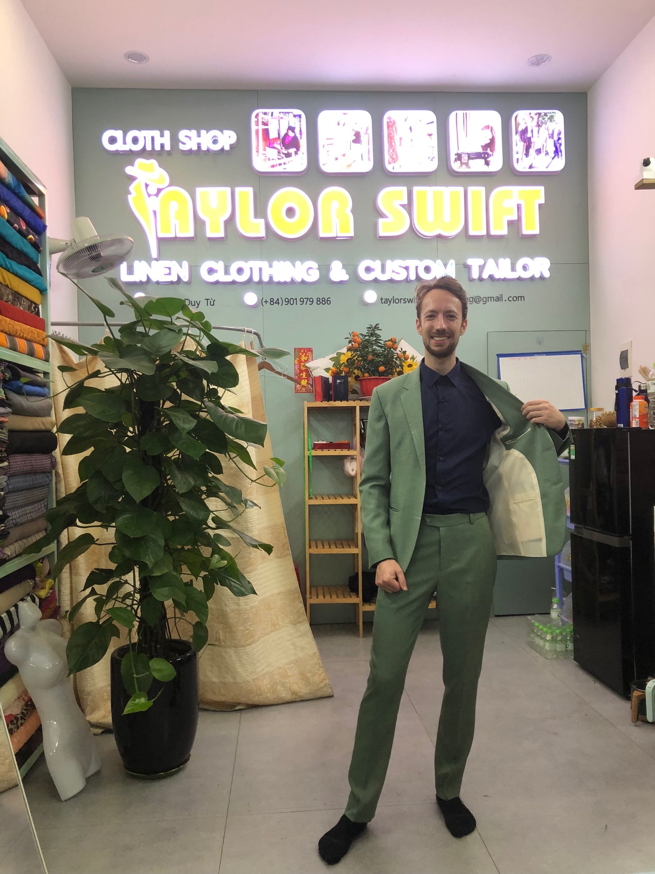 Man in a green suit poses inside a custom tailor shop with a "Taylor Swift" sign and rolls of fabric in the background.