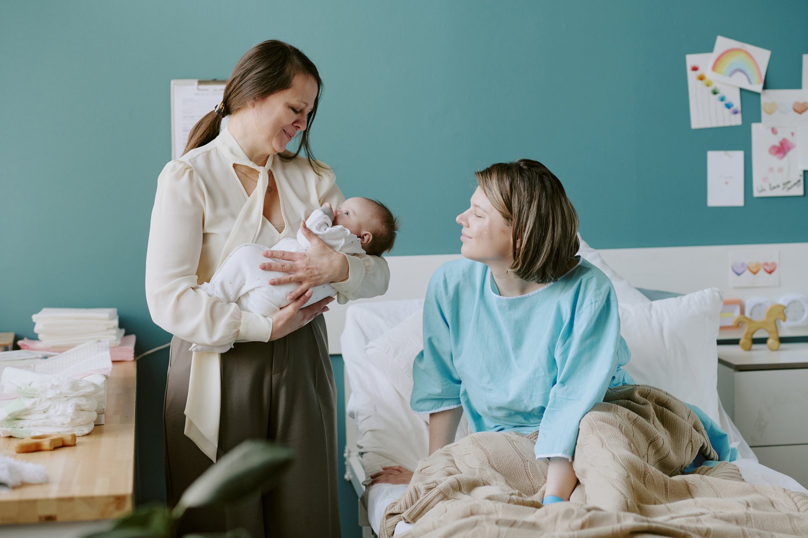 Grandmother holding newborn baby while new mother lying in hospital bed smiling at her. There are colorful drawings on wall in background