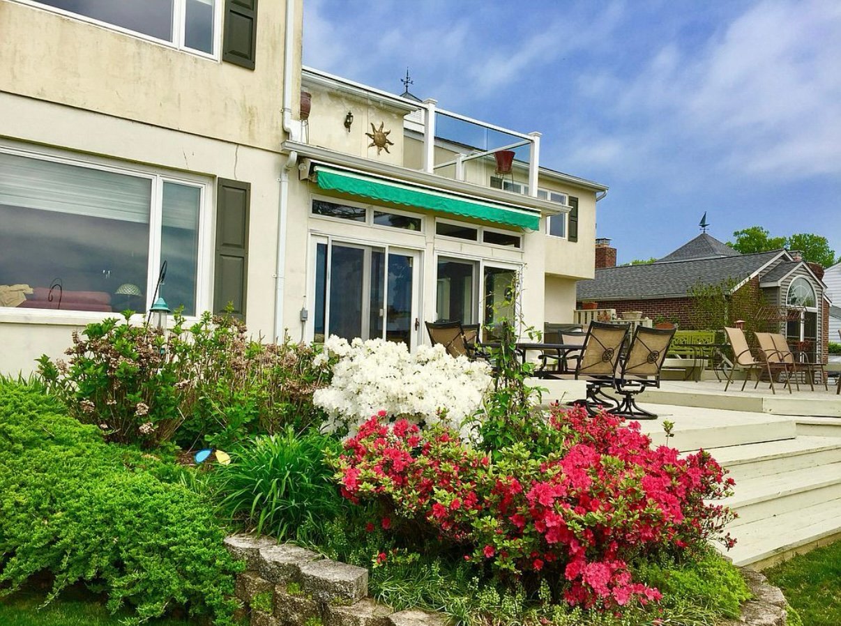 A two-story house with a garden, patio, outdoor seating, and flowering shrubs under a blue sky.
