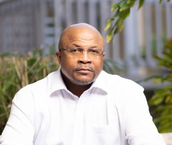 A man wearing glasses and a white shirt, seated outdoors against a blurred background of greenery and structures.