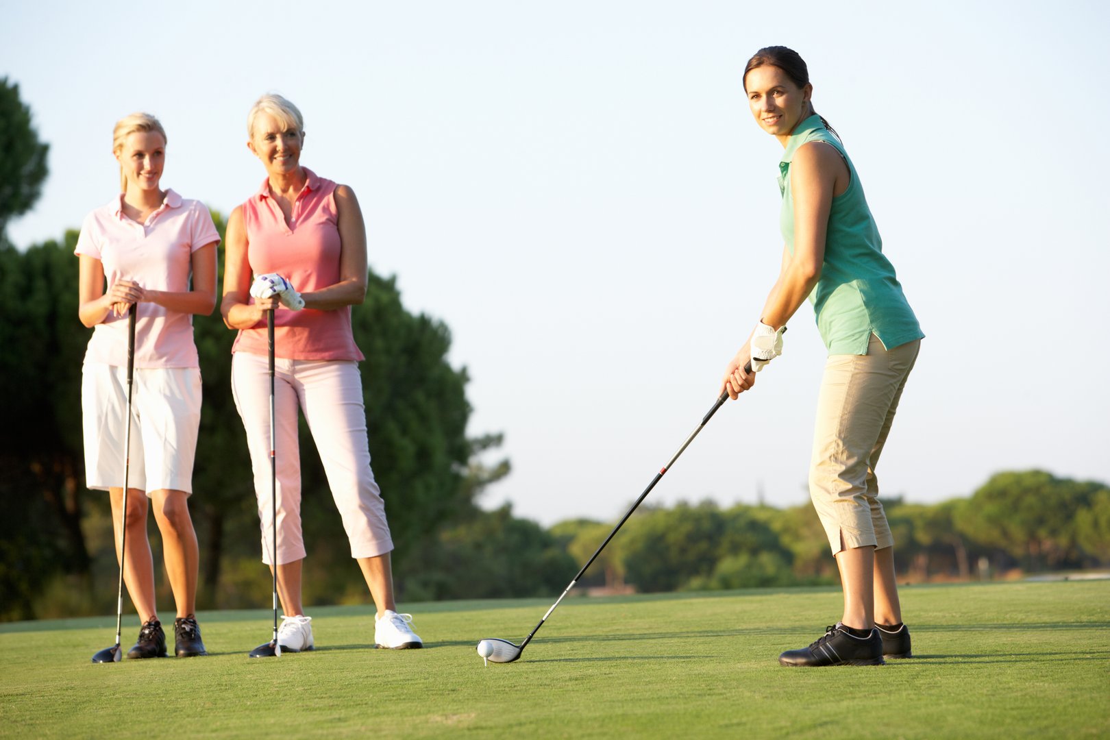 Three women in golf attire on a golf course, one preparing to swing while the other two watch and smile.