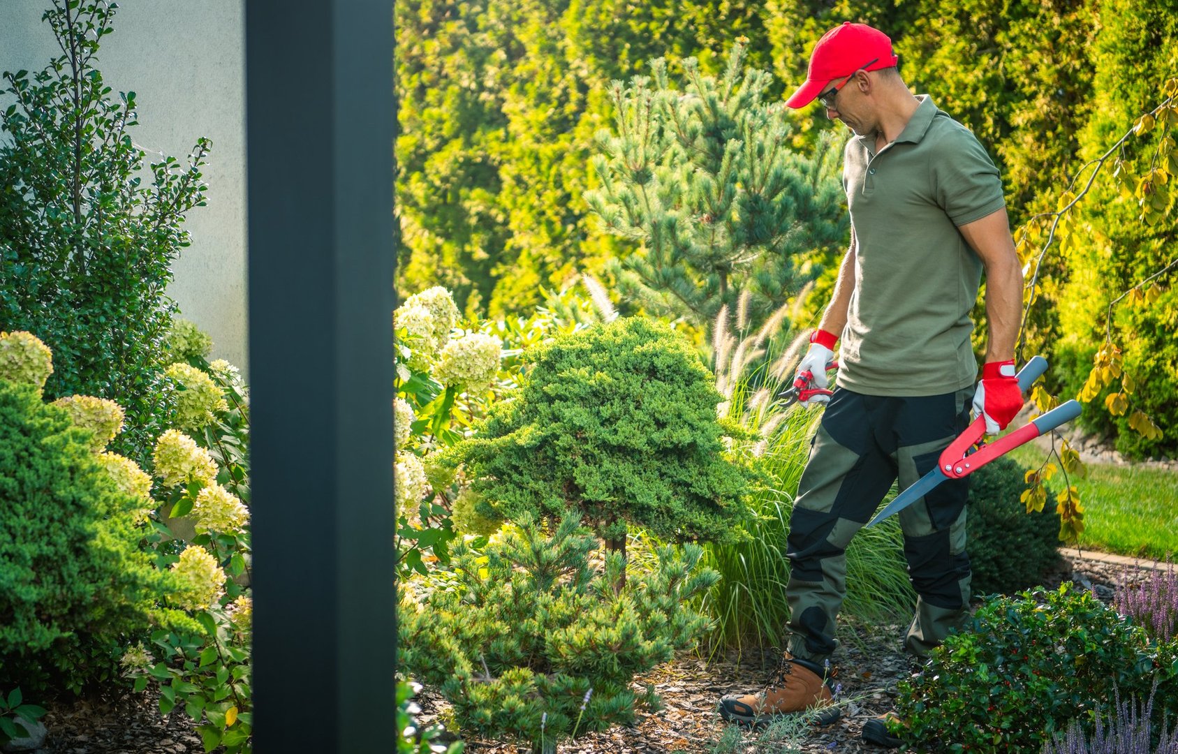 A gardener, wearing gloves and a cap, focuses on trimming bushes in a vibrant garden filled with various plants under bright sunlight.