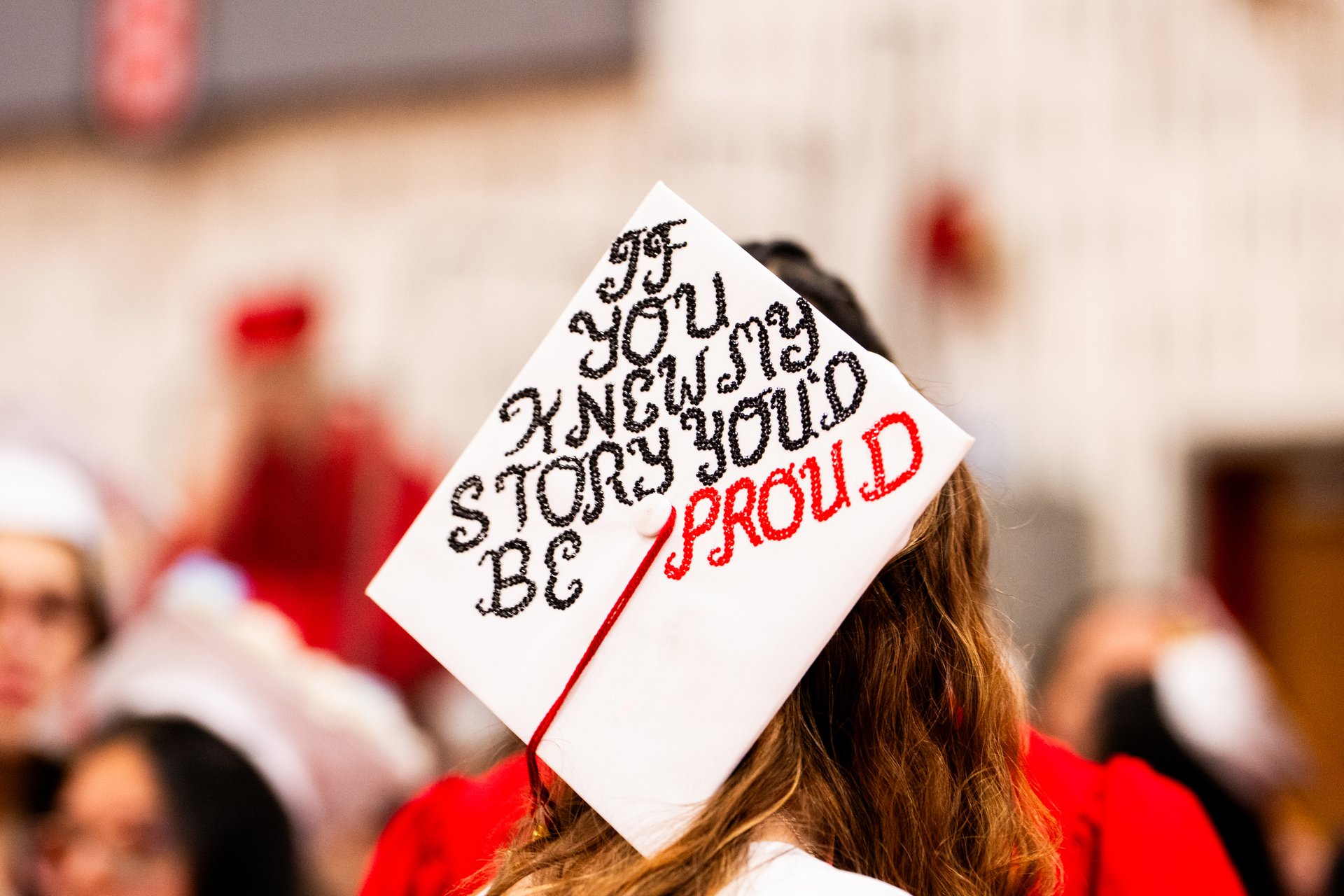 Graduate wearing white gown with decorated mortarboard cap that reads "If you knew my story, you would be proud." The unrecognizable teenager walks through a school gymnasium at graduation.