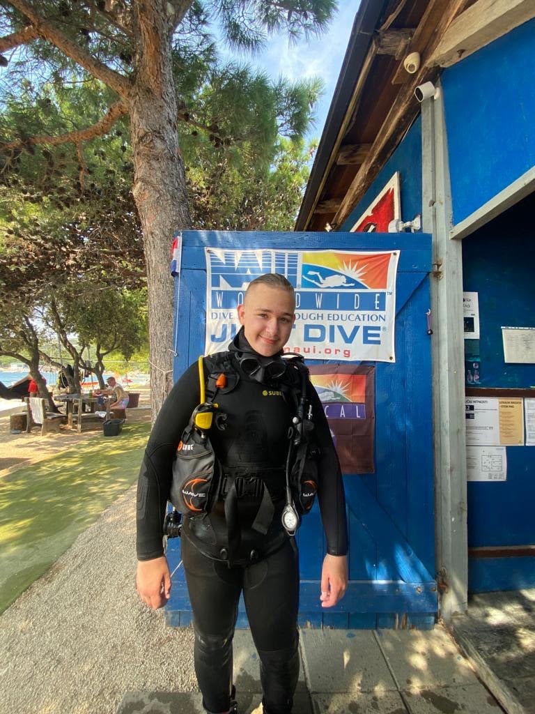 Scuba diver in a wetsuit standing outside a dive center with NAUI sign, surrounded by trees and outdoor seating area.