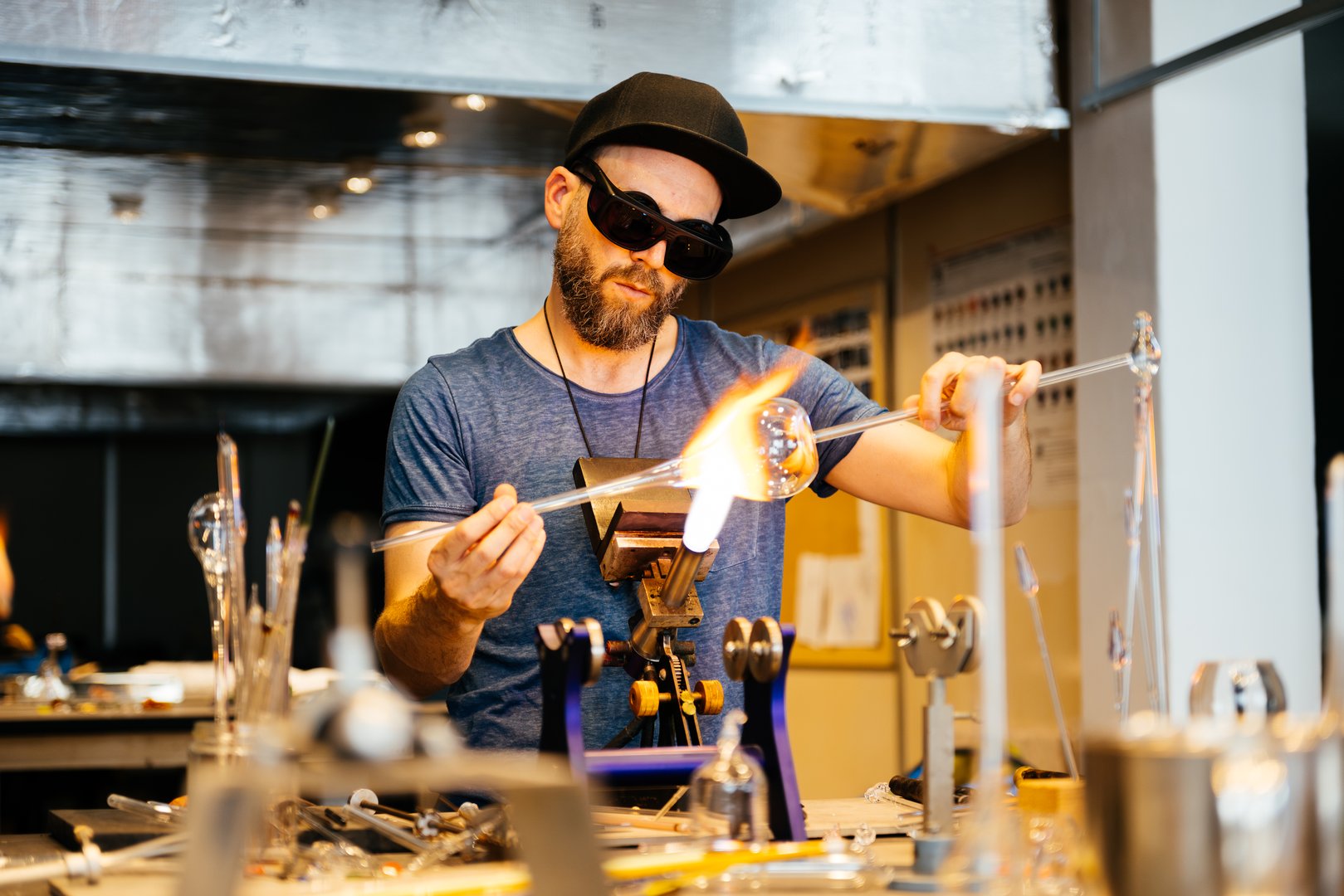 A glassblowing artisan works on a piece of glass, heating it with a torch and manipulating it with a long glassblowing rod. The artisan is wearing protective eyewear and a blue shirt.