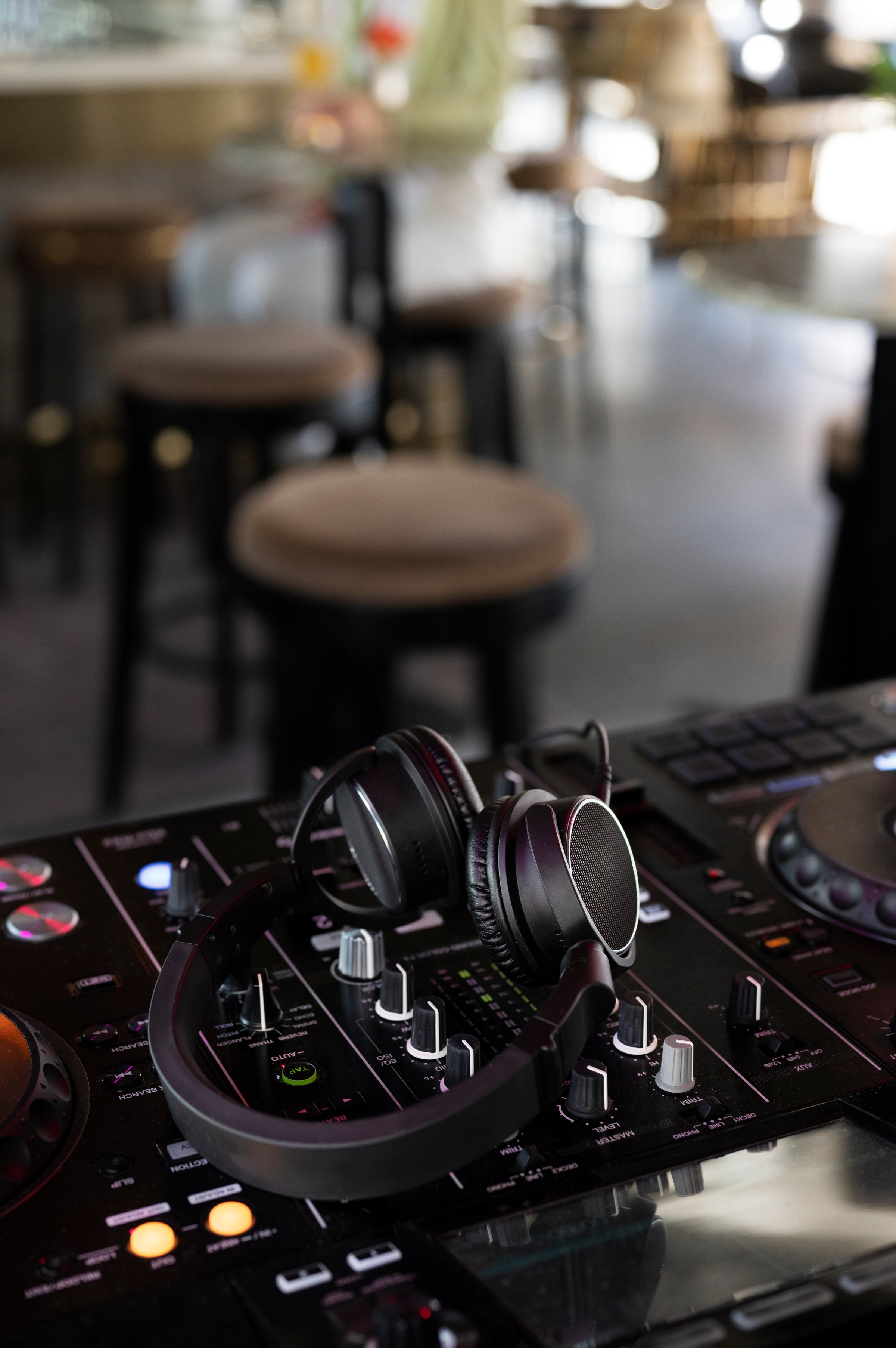 Close-up of DJ headphones on a mixing console in a dimly lit, modern cafe or bar setting with stools in the background.