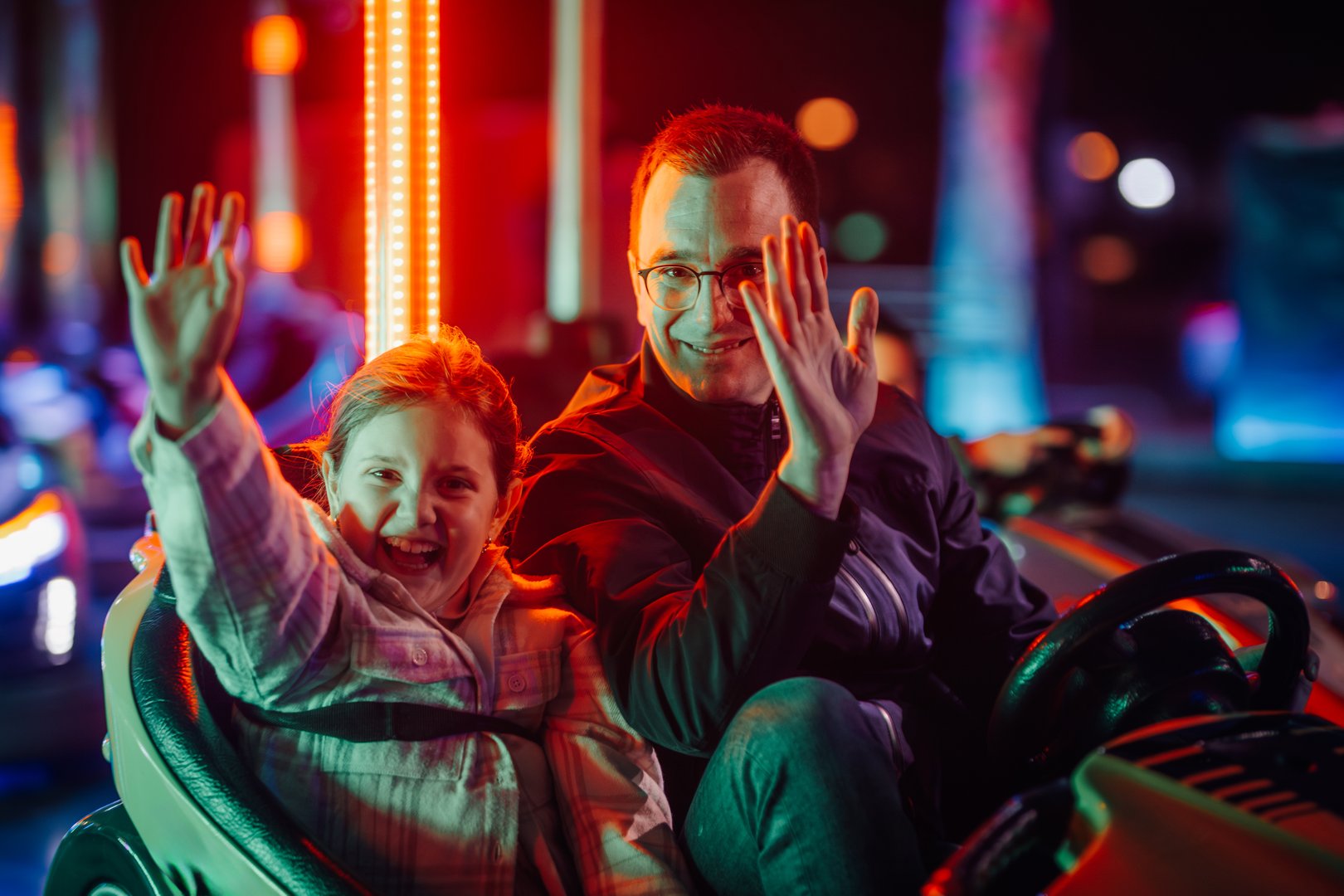 Father and daughter enjoying a fun night out, waving excitedly as they ride bumper cars at a brightly lit amusement park