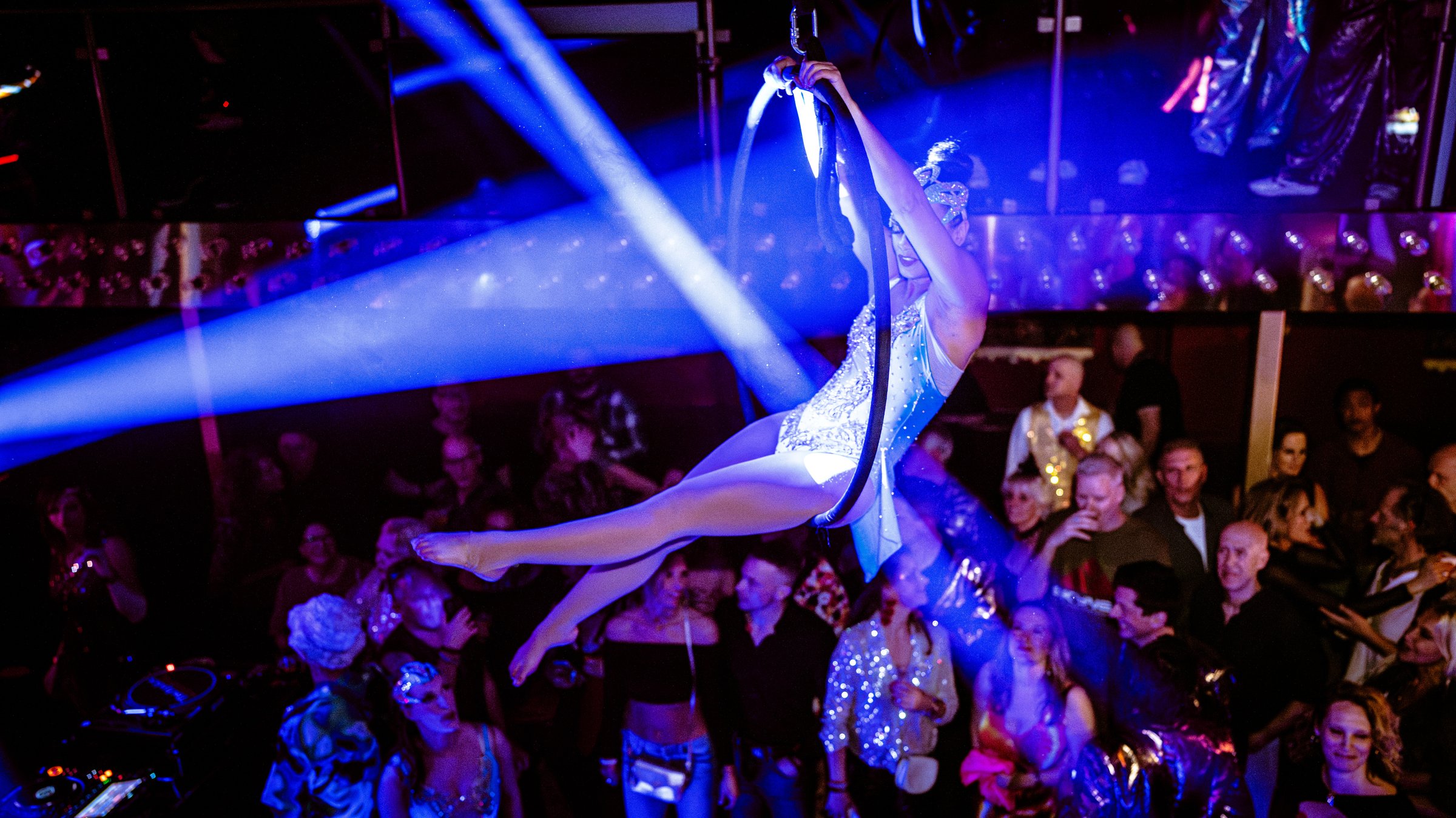 Aerial performer in a sparkly outfit swings above a crowded nightclub with colorful lights and audience watching.