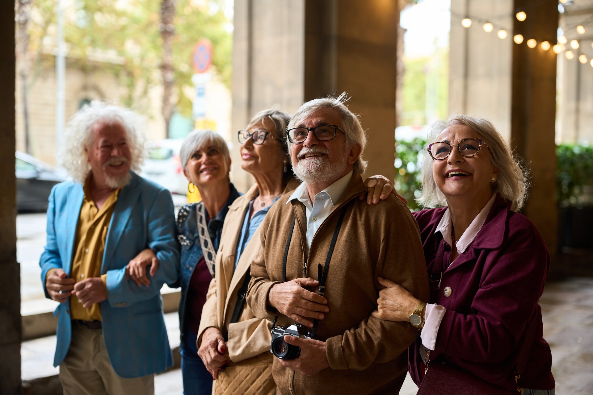 Group of happy senior adult friends exploring a city, smiling and looking up, arms linked during their travel