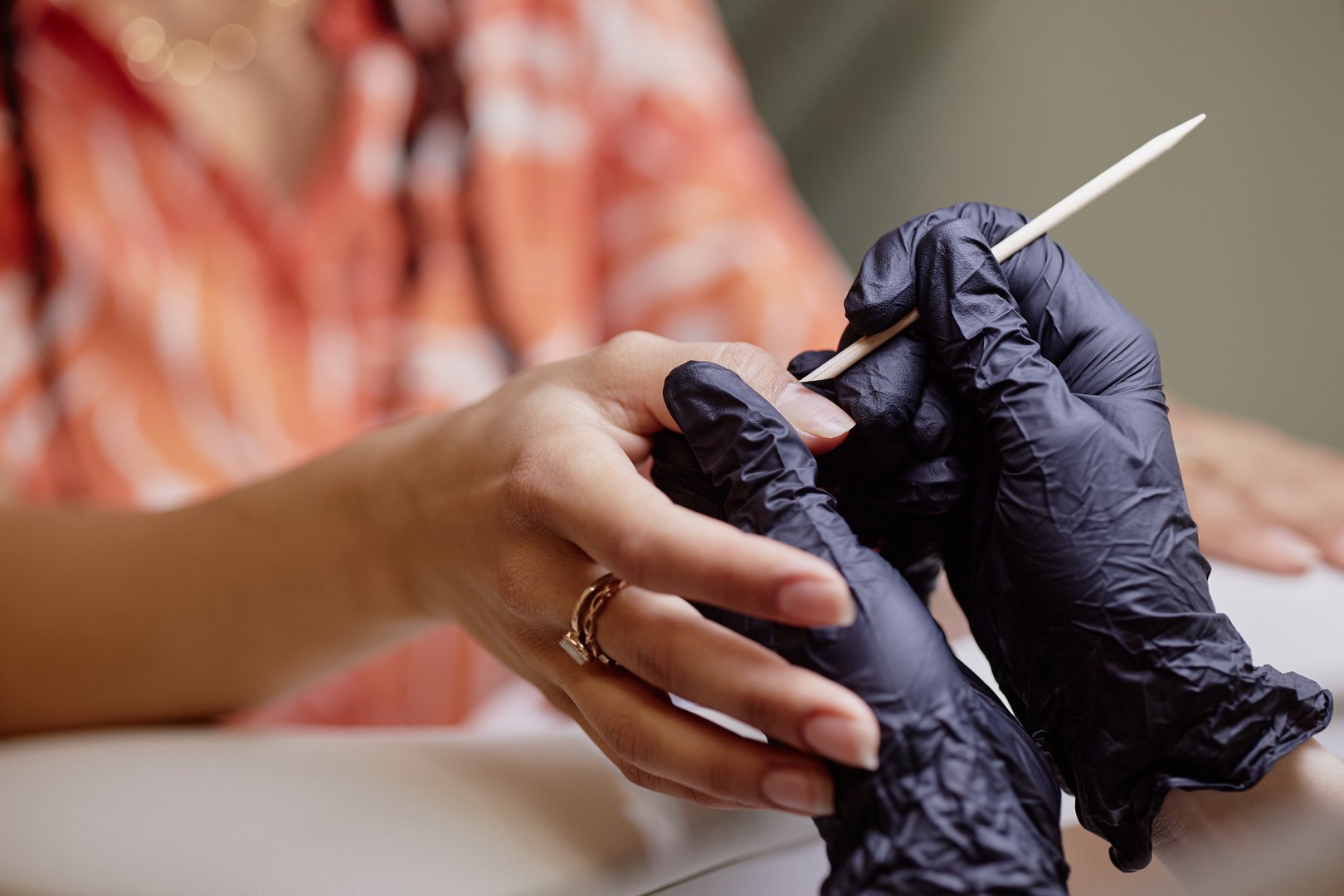 Biracial young adult woman receiving manicure from female professional wearing black gloves, hands visible while technician using wooden cuticle stick during nail care procedure
