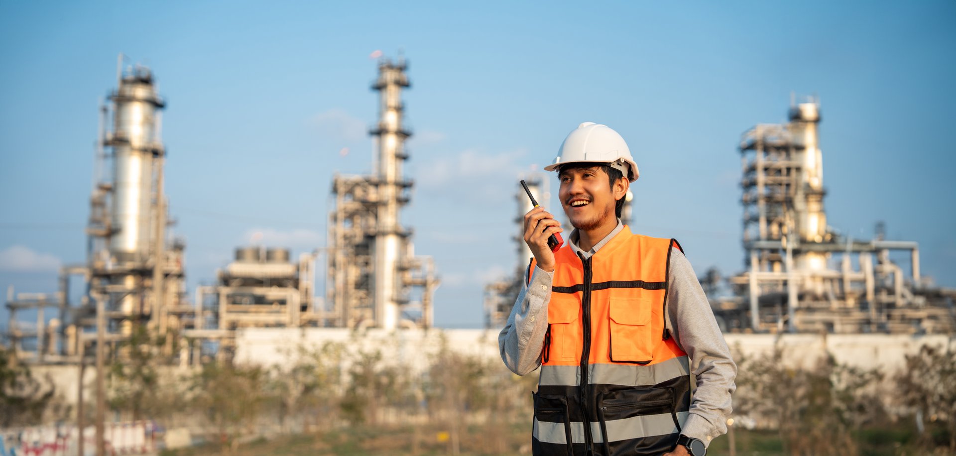 Asian engineer man with safety helmet standing front of oil refinery. Industry zone gas petrochemical. Factory oil storage tank and pipeline. Workers work in the refinery construction building