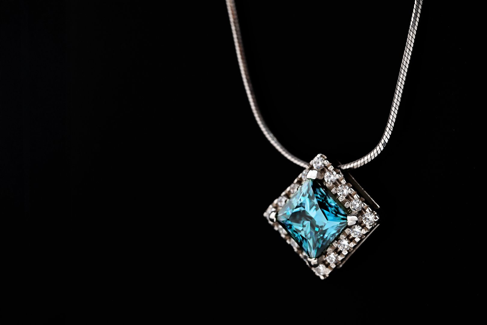A blue and white necklace sits on a black surface. The necklace is made of silver and has a diamond pendant. The necklace is simple and elegant, with a modern design