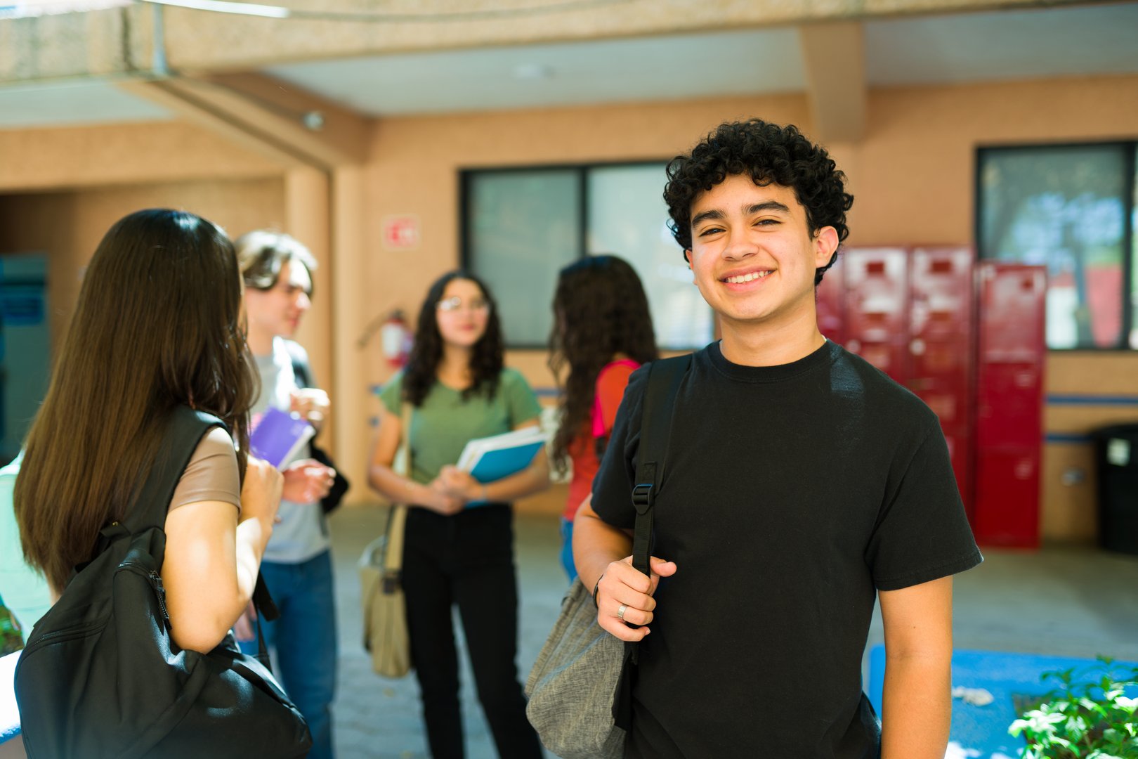 Happy latin high school student smiling in school hallway with classmates chatting in the background