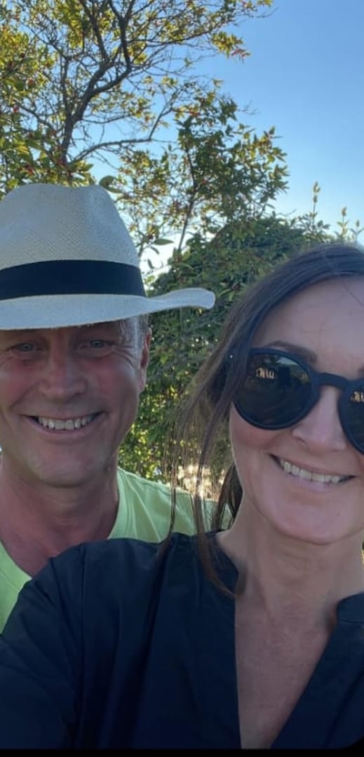 A man with a hat and a woman wearing sunglasses smiling for a selfie outdoors with trees in the background.