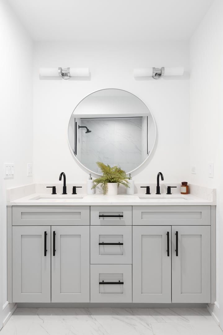 A renovated bathroom with a grey vanity cabinet, circular mirror with a view to a shower, and back faucets